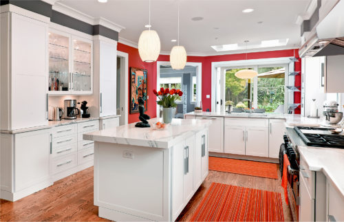 White shaker doors and stainless steel appliances highlight the grey veins on the white stone of this kitchen’s backsplash and countertops. Glass doors showcase the stemware and a built-in kitchen desk is tucked elegantly in a corner. Striking bar-style handles and pulls emphasize the strong lines of the kitchen’s design, keeping the style solidly contemporary.