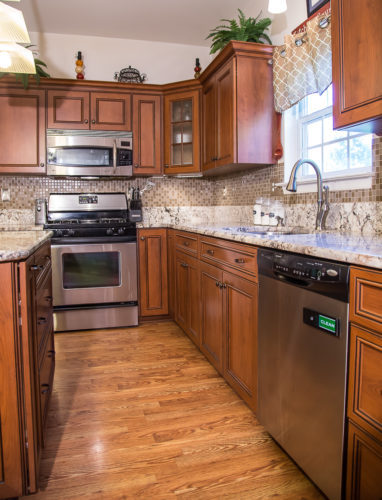 This kitchen was expanded and refaced in a warm wood-look laminate, and looks great! Existing cabinets were refaced and repaired, and new cabinets were added along with the new exteriors. Two of the kitchen cabinets received storage upgrades with full extension rollouts, while another got a pull out base organizer and a tip out tray added storage to the sink area. A classy and custom look was created by adding details like a mullion glass door, crown molding, a light rail and base molding.